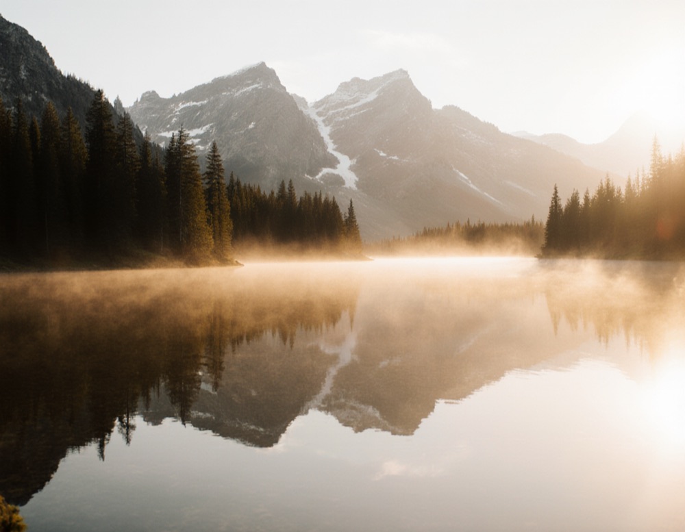 A misty mountain lake at sunrise with pine trees and snowy peaks reflected in the water